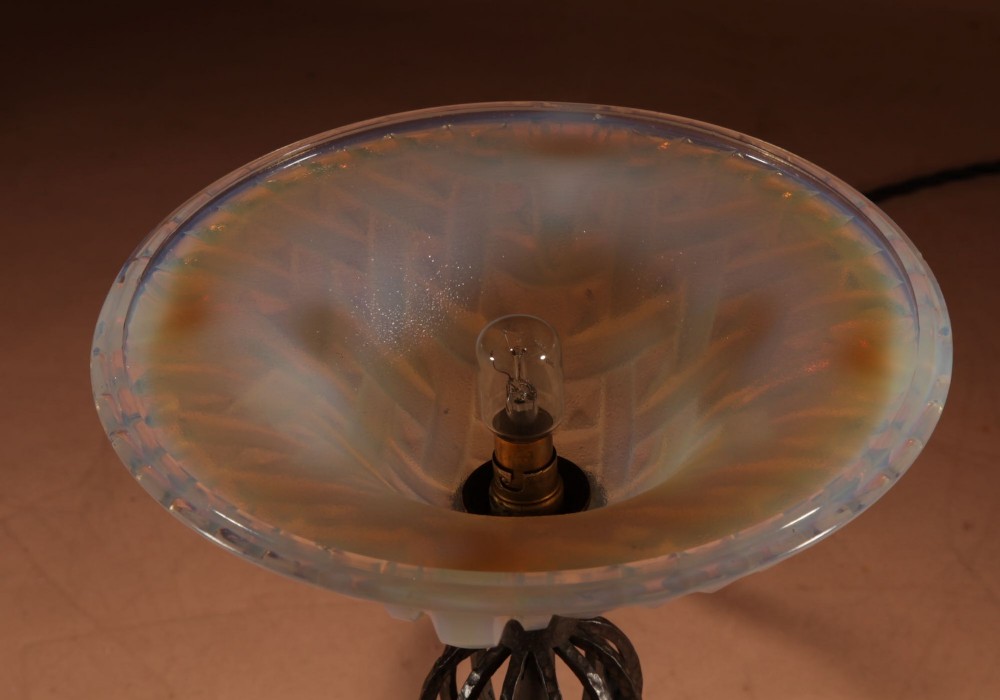 An illuminated French Art Deco table lamp with a flared opalescent glass shade set atop a dark, hammered wrought iron stem and circular base. View X