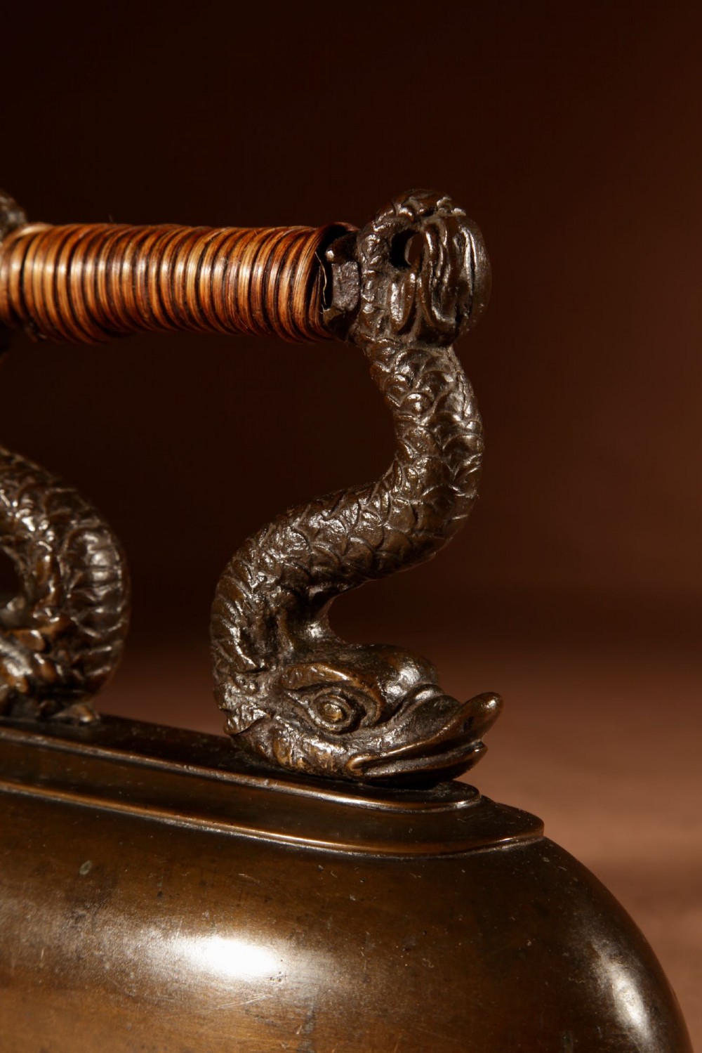A nineteenth-century French bronze flat iron featuring twin dolphin-shaped supports, a cane-wrapped handle, and a rear door mechanism. View VIII