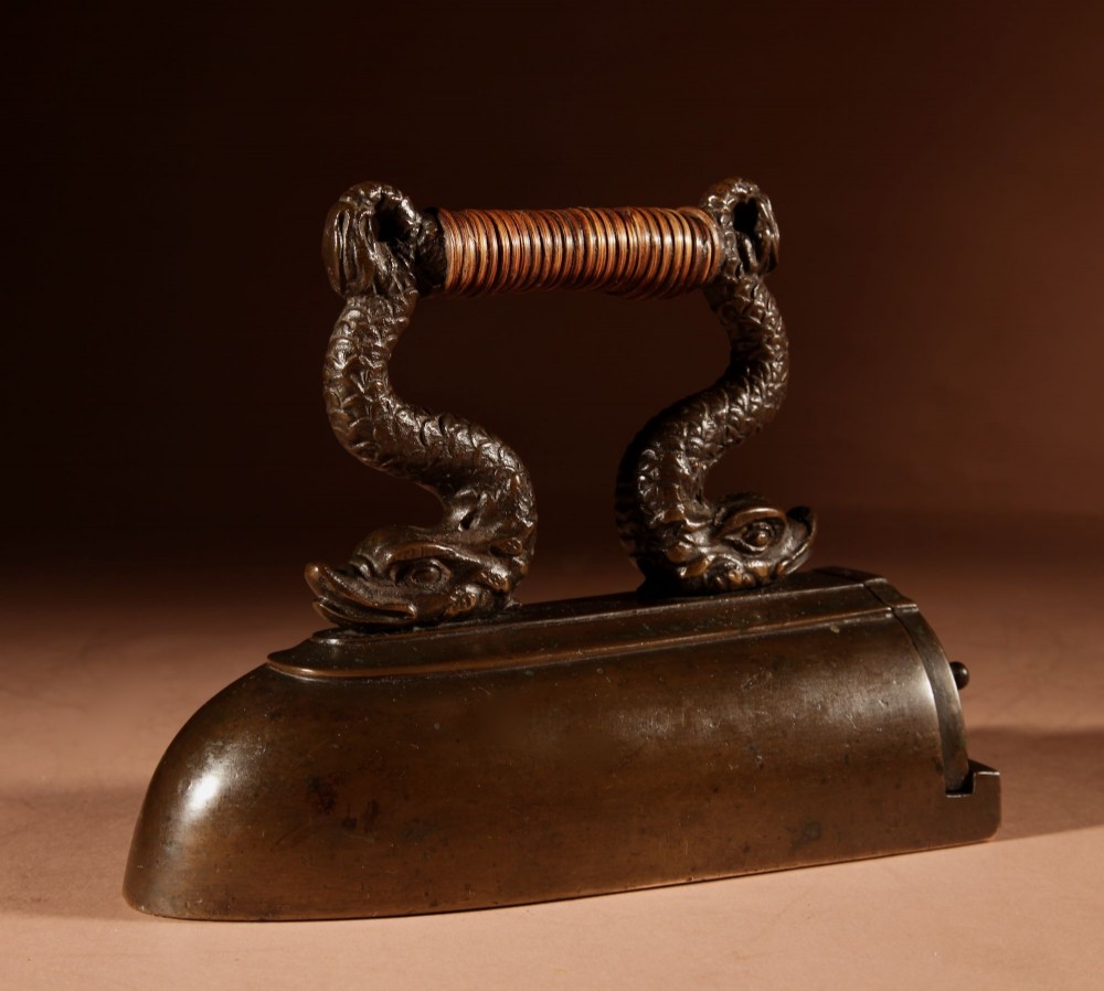 A nineteenth-century French bronze flat iron featuring twin dolphin-shaped supports, a cane-wrapped handle, and a rear door mechanism. View IX