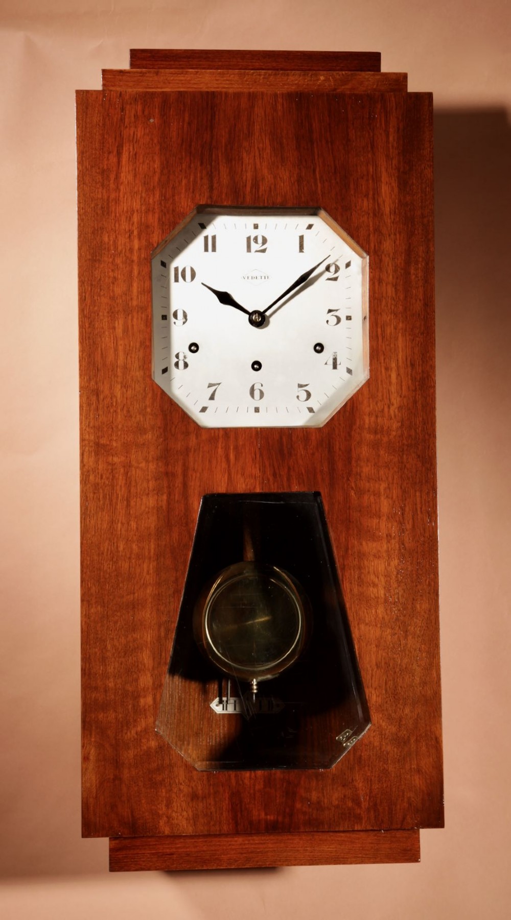 An Art Deco French walnut wall clock featuring a stepped top, an octagonal white dial face with Arabic numerals, and a visible brass pendulum bob behind a trapezoidal glass aperture. View II