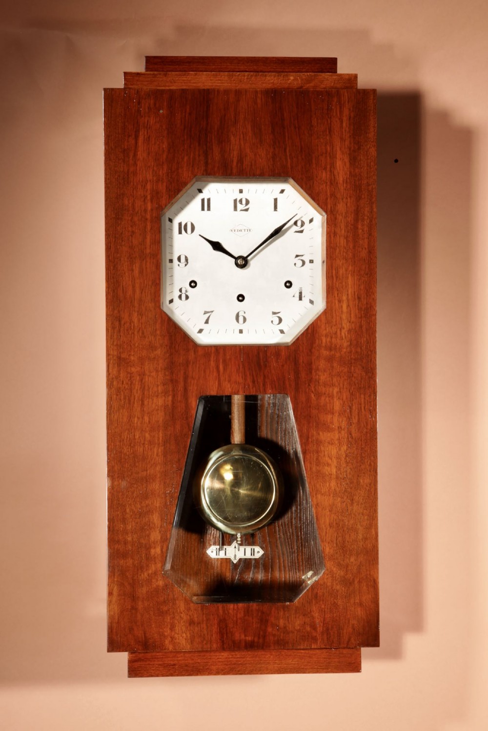 An Art Deco French walnut wall clock featuring a stepped top, an octagonal white dial face with Arabic numerals, and a visible brass pendulum bob behind a trapezoidal glass aperture.