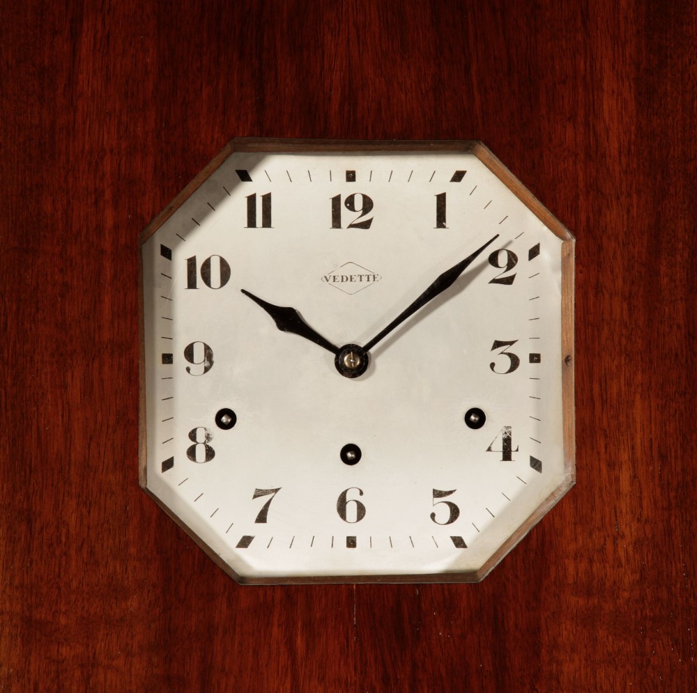 An Art Deco French walnut wall clock featuring a stepped top, an octagonal white dial face with Arabic numerals, and a visible brass pendulum bob behind a trapezoidal glass aperture. View III
