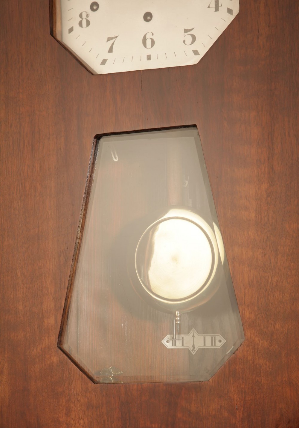 An Art Deco French walnut wall clock featuring a stepped top, an octagonal white dial face with Arabic numerals, and a visible brass pendulum bob behind a trapezoidal glass aperture. View IV