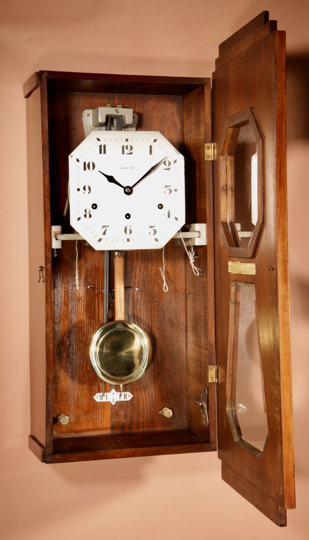 An Art Deco French walnut wall clock featuring a stepped top, an octagonal white dial face with Arabic numerals, and a visible brass pendulum bob behind a trapezoidal glass aperture. View V