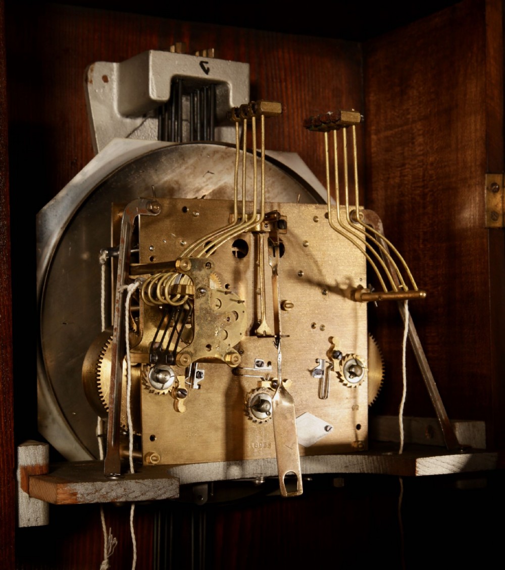 An Art Deco French walnut wall clock featuring a stepped top, an octagonal white dial face with Arabic numerals, and a visible brass pendulum bob behind a trapezoidal glass aperture. View VII