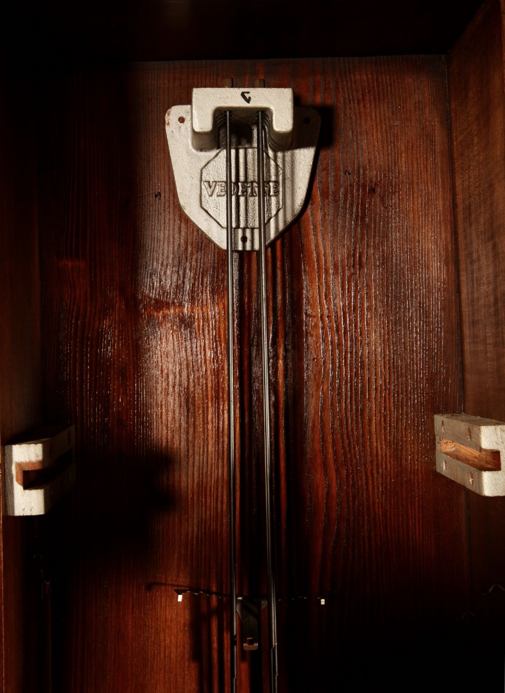 An Art Deco French walnut wall clock featuring a stepped top, an octagonal white dial face with Arabic numerals, and a visible brass pendulum bob behind a trapezoidal glass aperture. View VIII