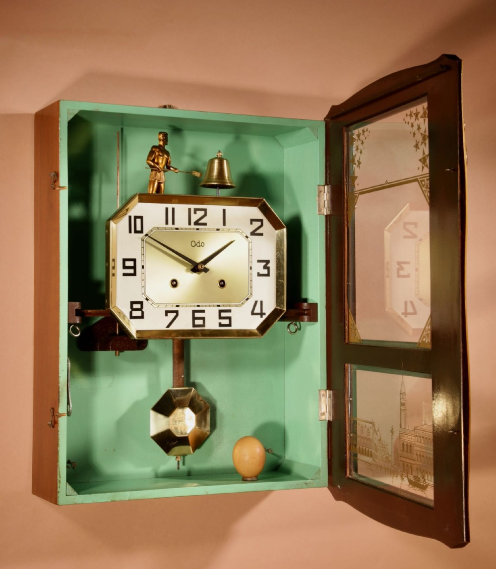 An Art Deco carved walnut wall clock by Odo featuring a brass automaton bell striker and a Venetian glass lower window. View X