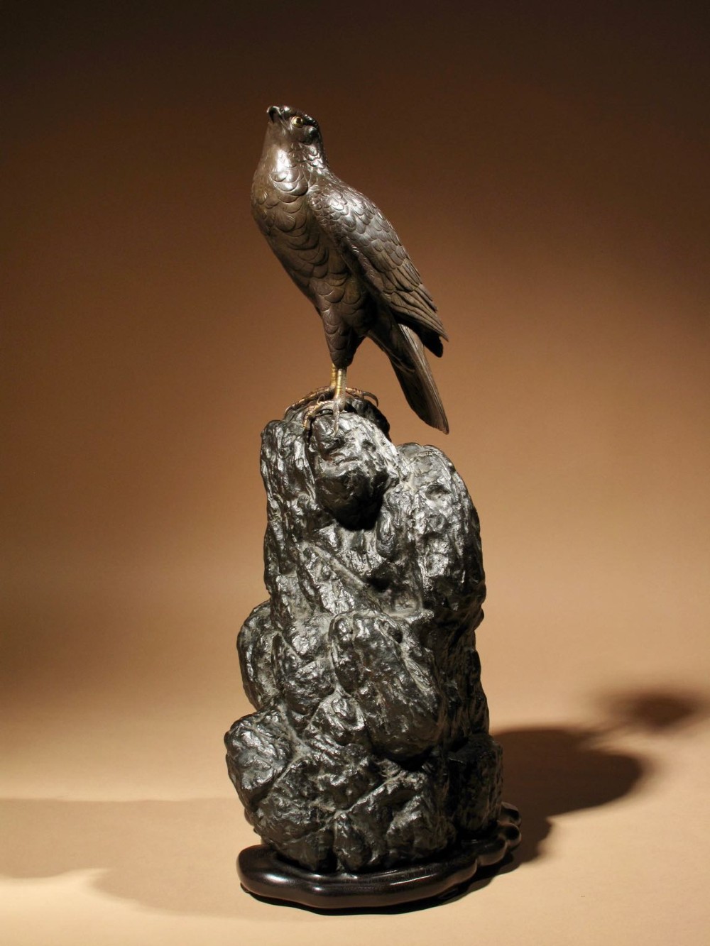 A patinated bronze sculpture depicting a hawk with detailed feather chasing perched atop a textured rocky outcrop, mounted on a carved wooden base. View II