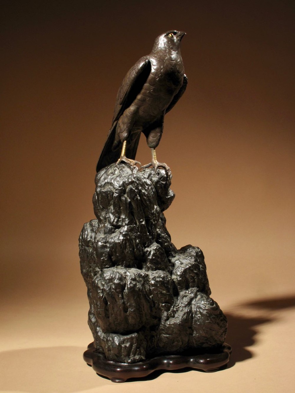 A patinated bronze sculpture depicting a hawk with detailed feather chasing perched atop a textured rocky outcrop, mounted on a carved wooden base.