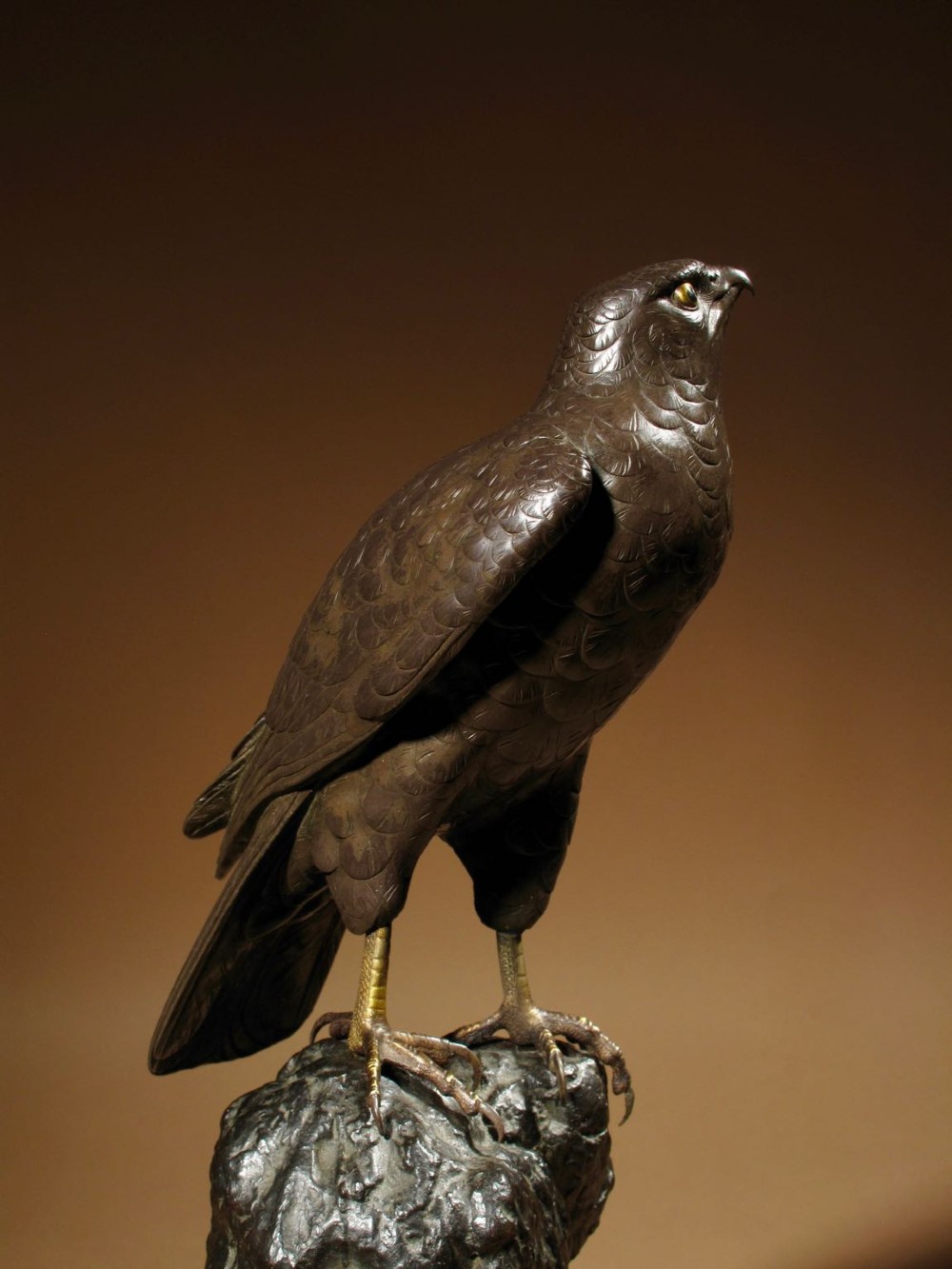 A patinated bronze sculpture depicting a hawk with detailed feather chasing perched atop a textured rocky outcrop, mounted on a carved wooden base. View III