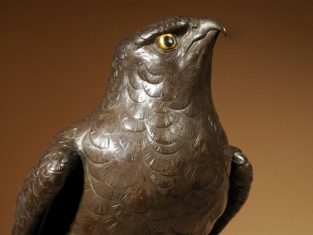 A patinated bronze sculpture depicting a hawk with detailed feather chasing perched atop a textured rocky outcrop, mounted on a carved wooden base. View V