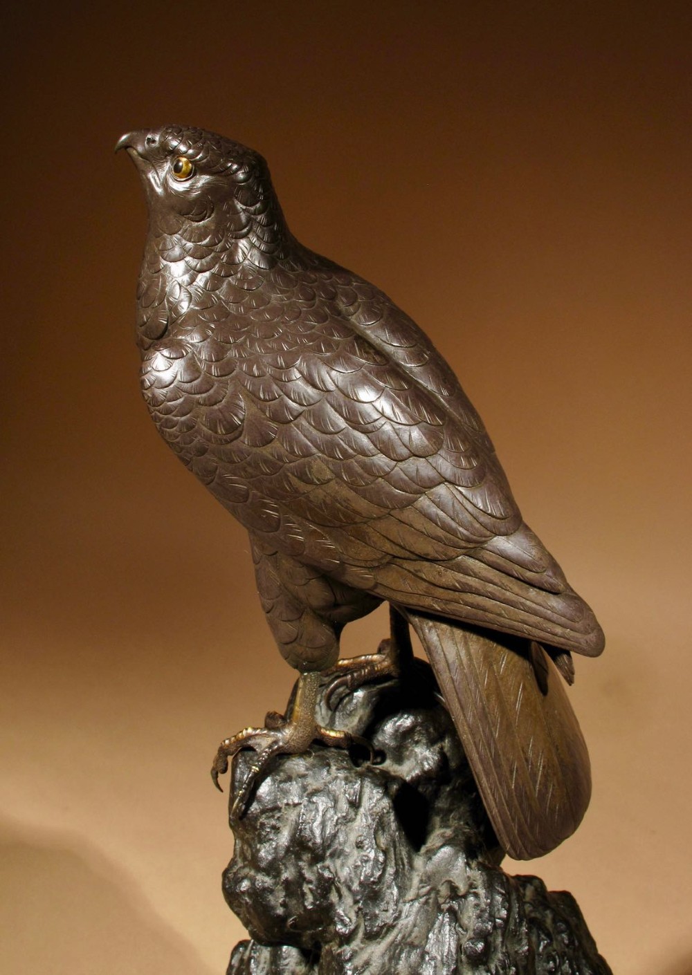 A patinated bronze sculpture depicting a hawk with detailed feather chasing perched atop a textured rocky outcrop, mounted on a carved wooden base. View VI