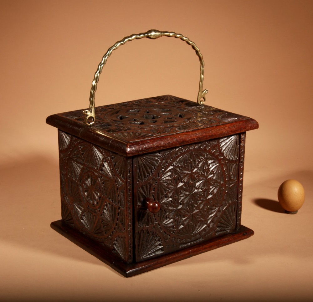 A dark carved oak foot stove featuring geometric chip carving, a pierced top lid, a hinged side door, and a twisted brass carrying handle.