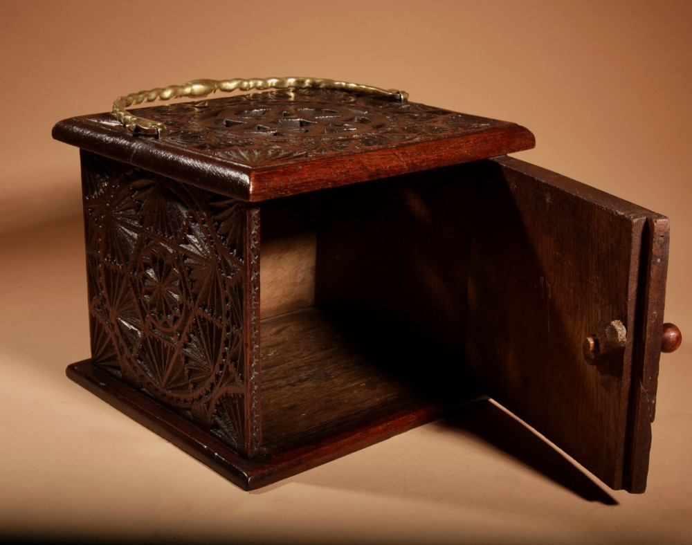 A dark carved oak foot stove featuring geometric chip carving, a pierced top lid, a hinged side door, and a twisted brass carrying handle. View V