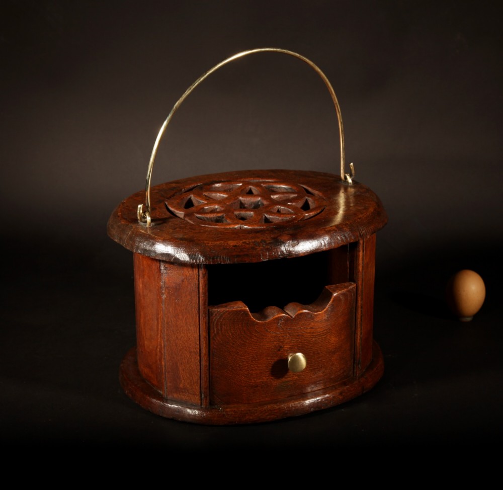 An oval dark oak foot stove featuring a carved geometric top aperture and an arched brass carrying handle.