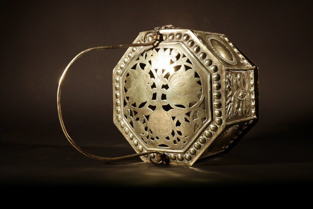 An 18th-century octagonal brass foot stove featuring hand-embossed floral panels, a pierced lid, and an overhead carrying handle. View V