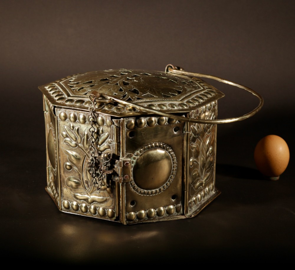 An 18th-century octagonal brass foot stove featuring hand-embossed floral panels, a pierced lid, and an overhead carrying handle.