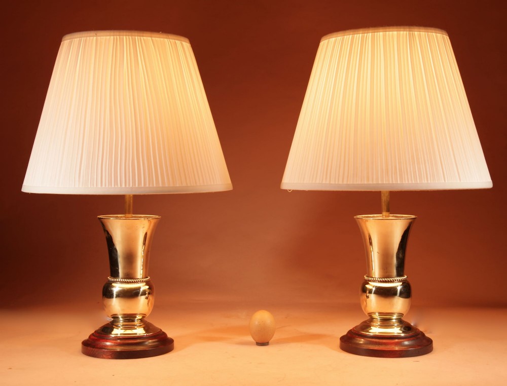 A pair of polished brass table lamps with flared bases on dark wooden plinths, topped with cream pleated shades.