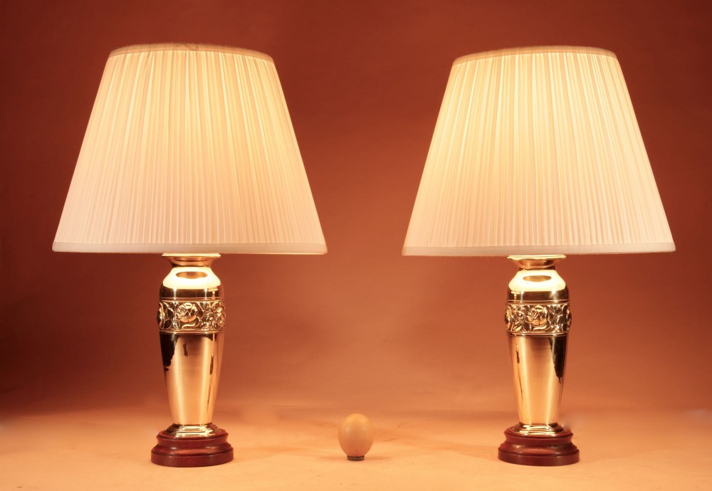 A pair of French Art Deco polished brass urn-form table lamps featuring embossed floral bands on wooden bases with pleated cream shades.