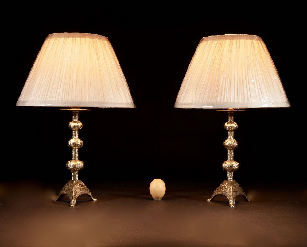 A pair of gold brass table lamps featuring relief-patterned shafts, tripod bases, and cream pleated shades.