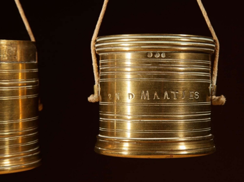 An 1846 Dutch brass bushel balance set featuring a balance beam, two cylindrical brass measuring buckets, calibrated brass weights, a wooden striker, and a striped fabric pouch. View V