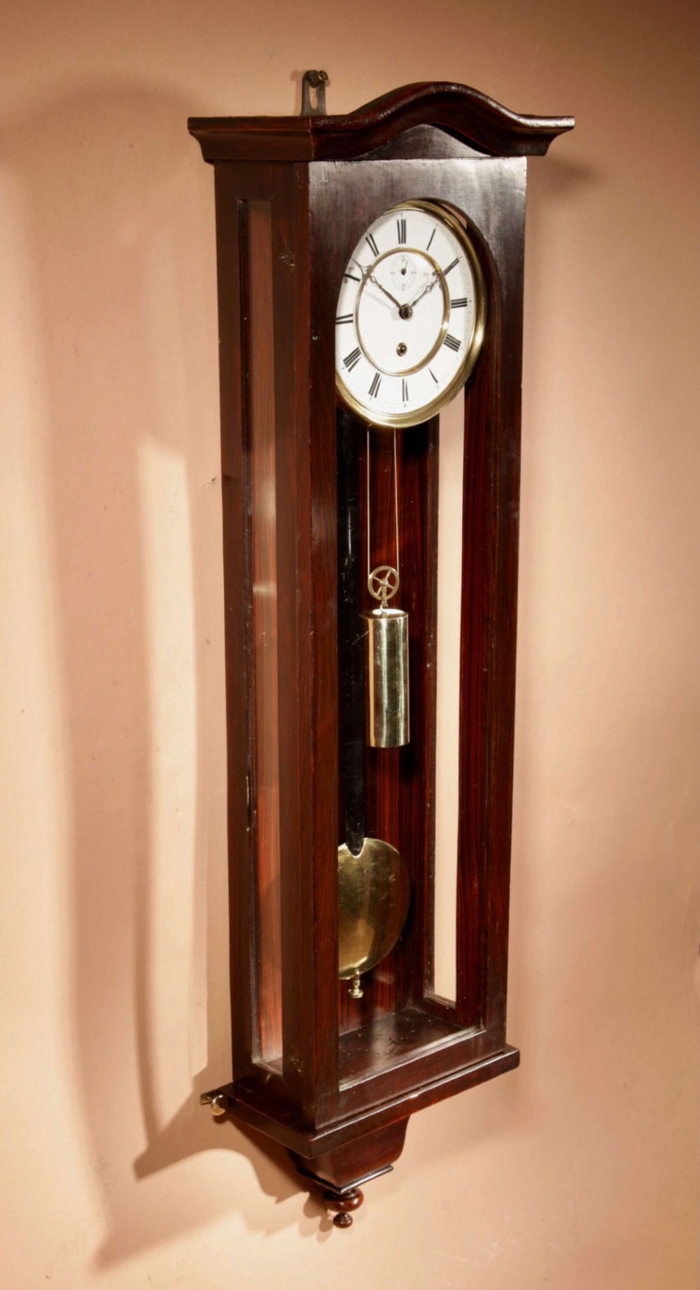 An antique dark wood Vienna regulator wall clock featuring a glazed case, white enamel dial with Roman numerals, and a polished brass pendulum. View IV