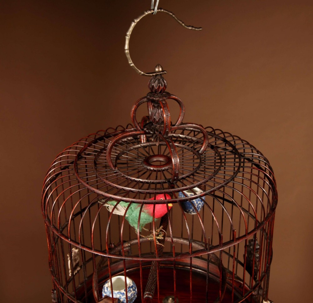 A cylindrical dark carved bamboo Chinese bird cage featuring a metal hanging hook, blue and white porcelain feeding cups, intricate carved plaques, and a matching circular wooden base. View III