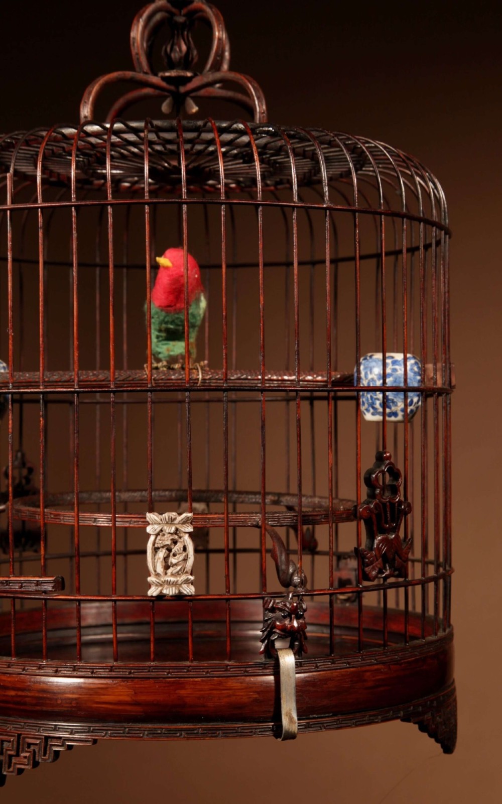 A cylindrical dark carved bamboo Chinese bird cage featuring a metal hanging hook, blue and white porcelain feeding cups, intricate carved plaques, and a matching circular wooden base. View IV