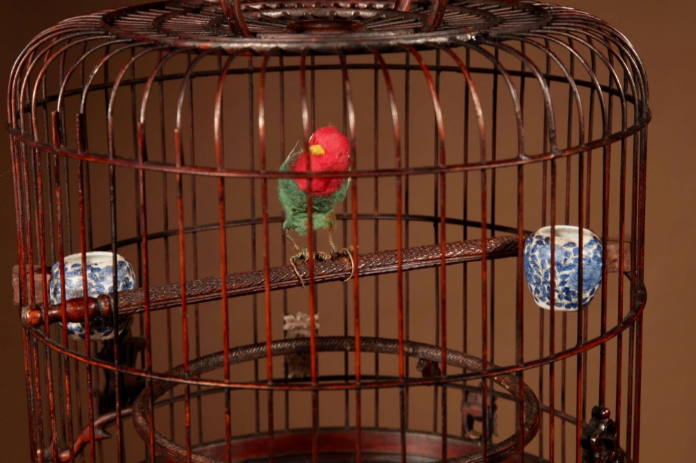 A cylindrical dark carved bamboo Chinese bird cage featuring a metal hanging hook, blue and white porcelain feeding cups, intricate carved plaques, and a matching circular wooden base. View V