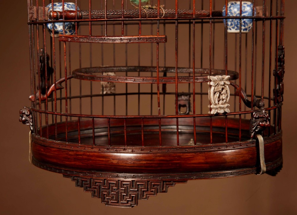 A cylindrical dark carved bamboo Chinese bird cage featuring a metal hanging hook, blue and white porcelain feeding cups, intricate carved plaques, and a matching circular wooden base. View VI