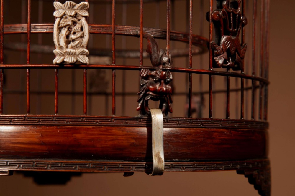 A cylindrical dark carved bamboo Chinese bird cage featuring a metal hanging hook, blue and white porcelain feeding cups, intricate carved plaques, and a matching circular wooden base. View VIII