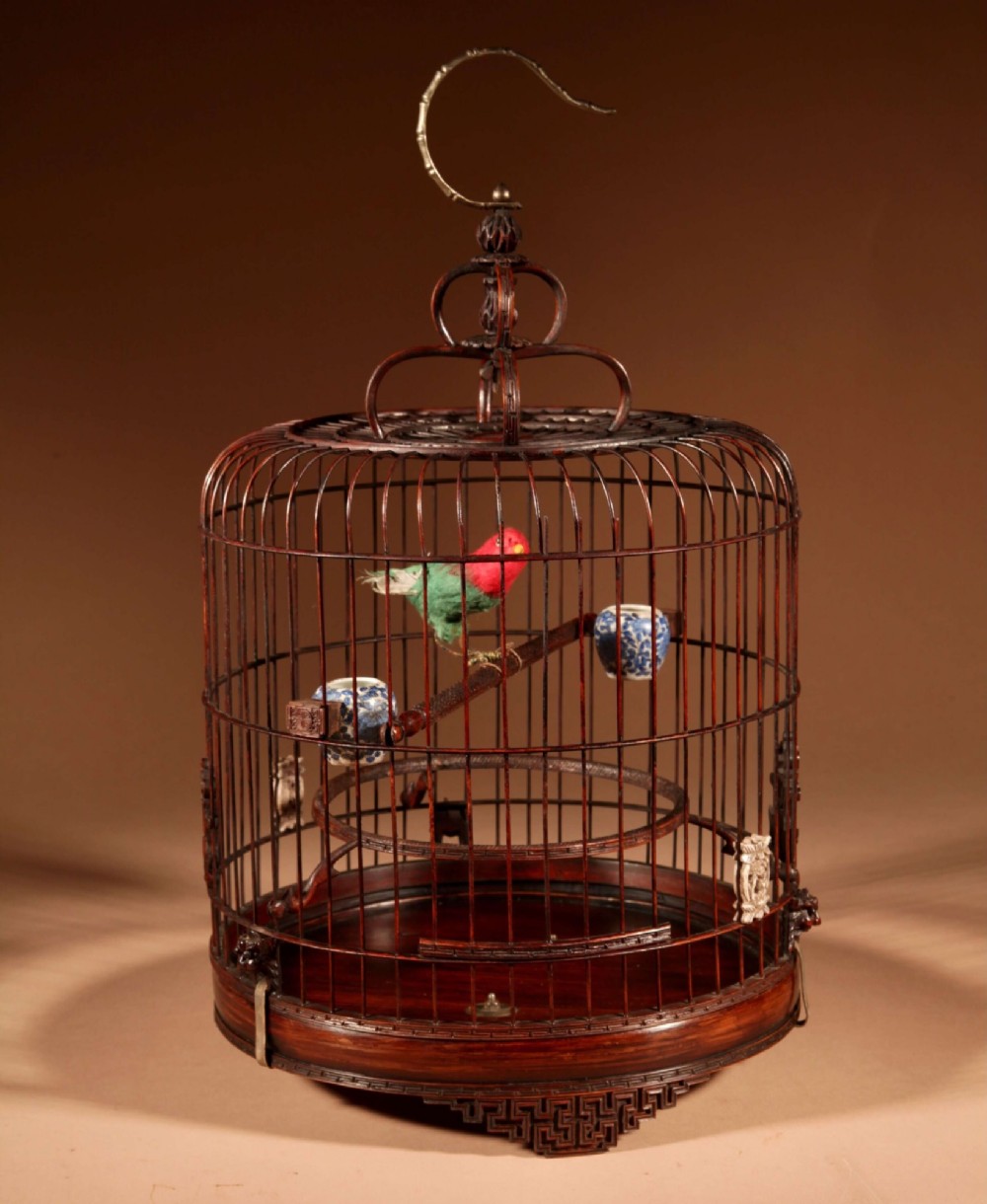 A cylindrical dark carved bamboo Chinese bird cage featuring a metal hanging hook, blue and white porcelain feeding cups, intricate carved plaques, and a matching circular wooden base. View IX