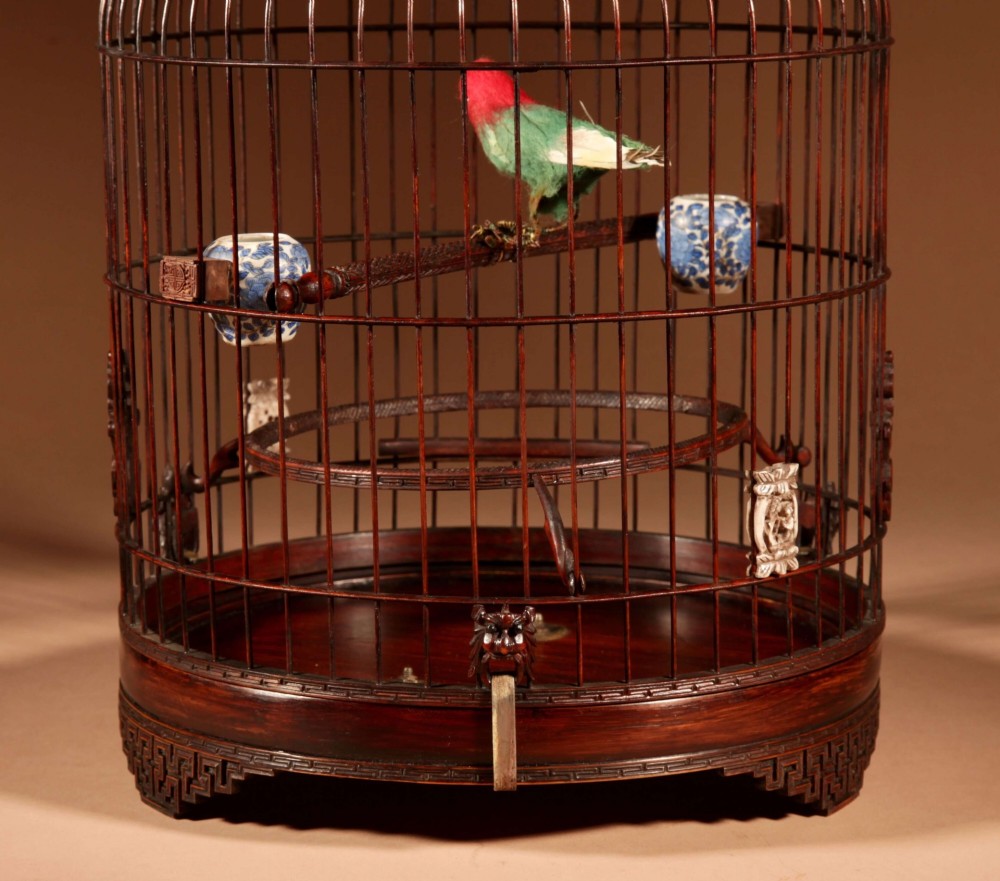 A cylindrical dark carved bamboo Chinese bird cage featuring a metal hanging hook, blue and white porcelain feeding cups, intricate carved plaques, and a matching circular wooden base. View X