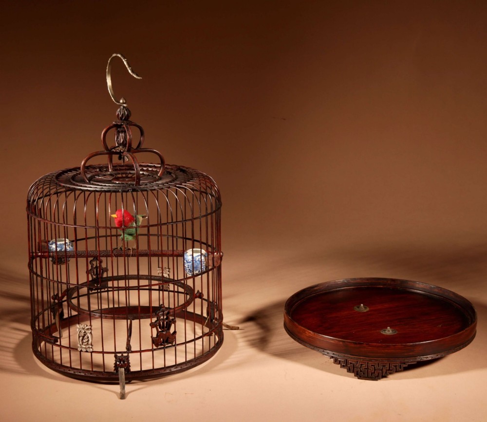 A cylindrical dark carved bamboo Chinese bird cage featuring a metal hanging hook, blue and white porcelain feeding cups, intricate carved plaques, and a matching circular wooden base.