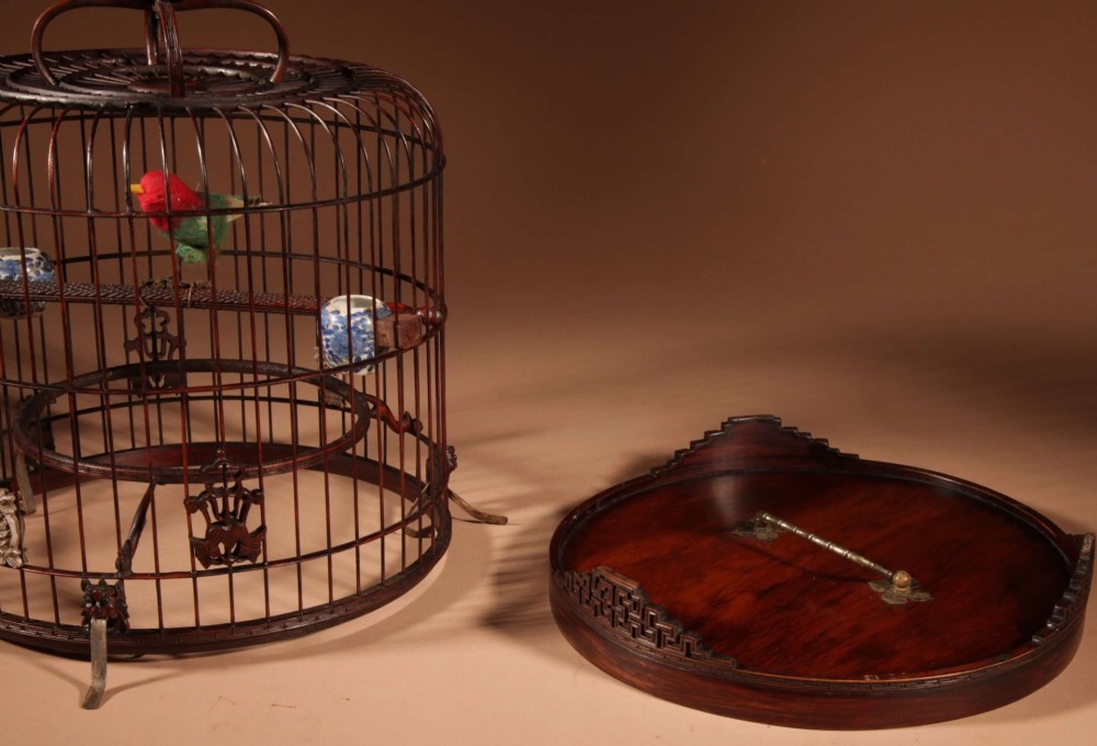 A cylindrical dark carved bamboo Chinese bird cage featuring a metal hanging hook, blue and white porcelain feeding cups, intricate carved plaques, and a matching circular wooden base. View XII
