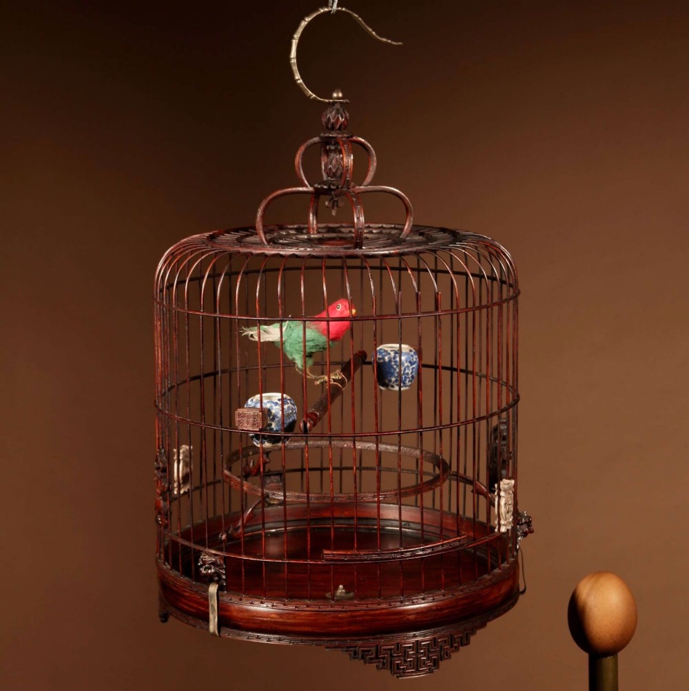 A cylindrical dark carved bamboo Chinese bird cage featuring a metal hanging hook, blue and white porcelain feeding cups, intricate carved plaques, and a matching circular wooden base. View XIII