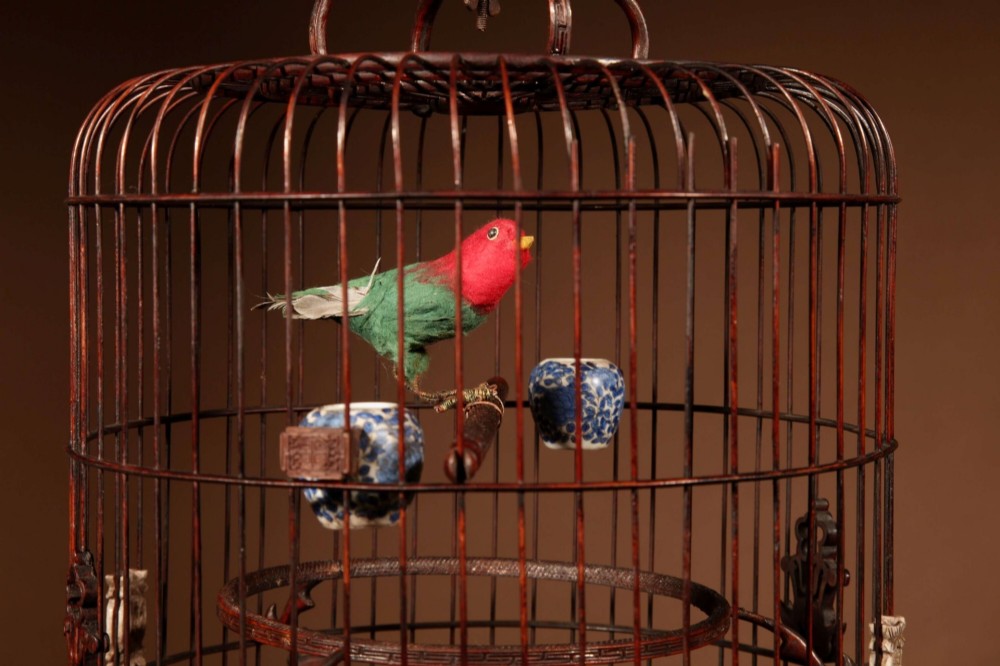 A cylindrical dark carved bamboo Chinese bird cage featuring a metal hanging hook, blue and white porcelain feeding cups, intricate carved plaques, and a matching circular wooden base. View XV