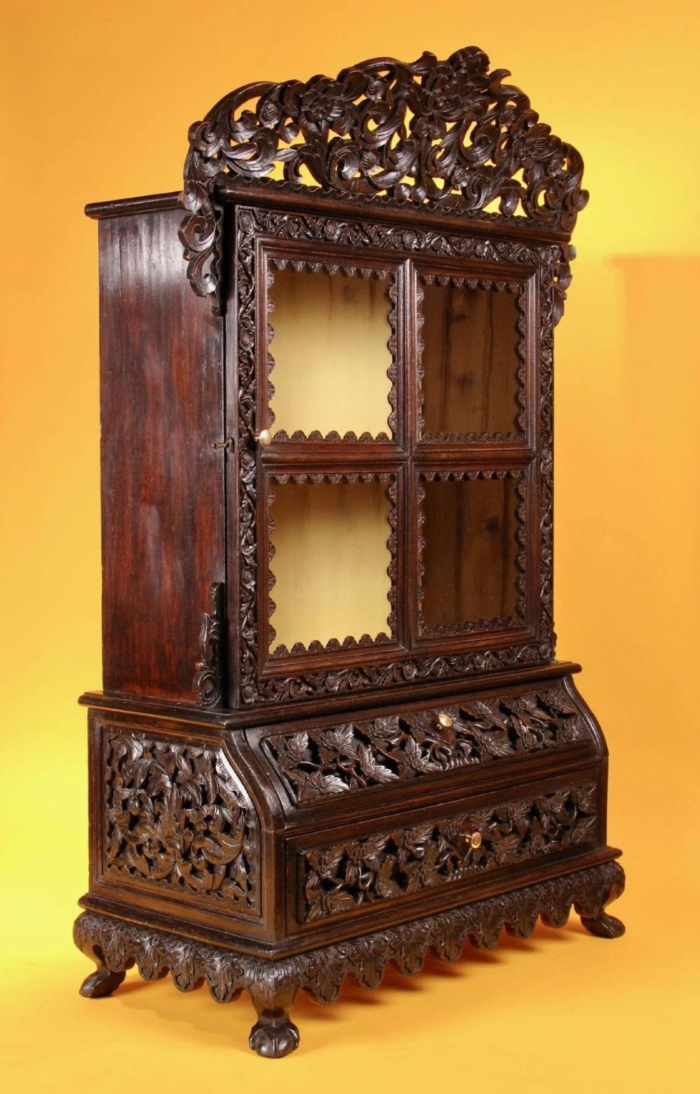 A 19th-century Anglo-Indian dark hardwood small display cabinet featuring intricate pierced foliate carvings, an arched pediment, a glazed four-pane door with scalloped moulding, and two carved drawers at the base. View III