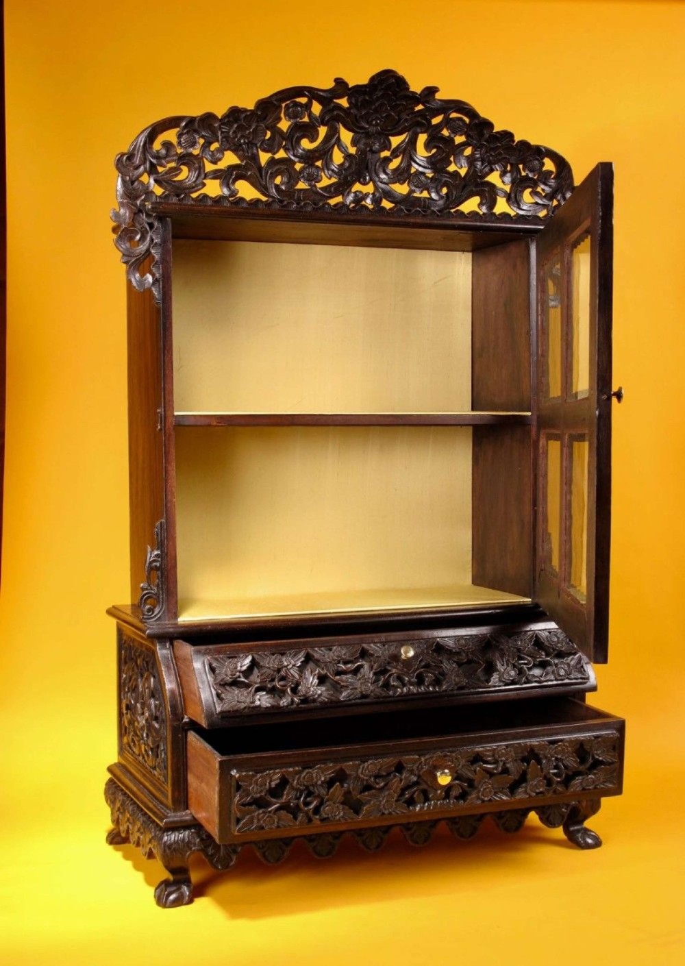 A 19th-century Anglo-Indian dark hardwood small display cabinet featuring intricate pierced foliate carvings, an arched pediment, a glazed four-pane door with scalloped moulding, and two carved drawers at the base. View V