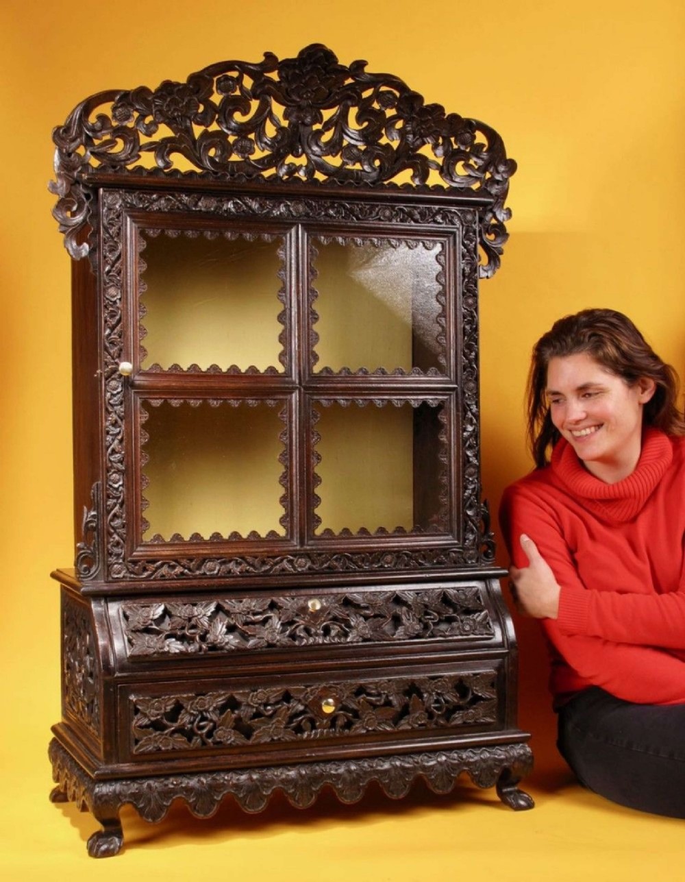 A 19th-century Anglo-Indian dark hardwood small display cabinet featuring intricate pierced foliate carvings, an arched pediment, a glazed four-pane door with scalloped moulding, and two carved drawers at the base. View IX