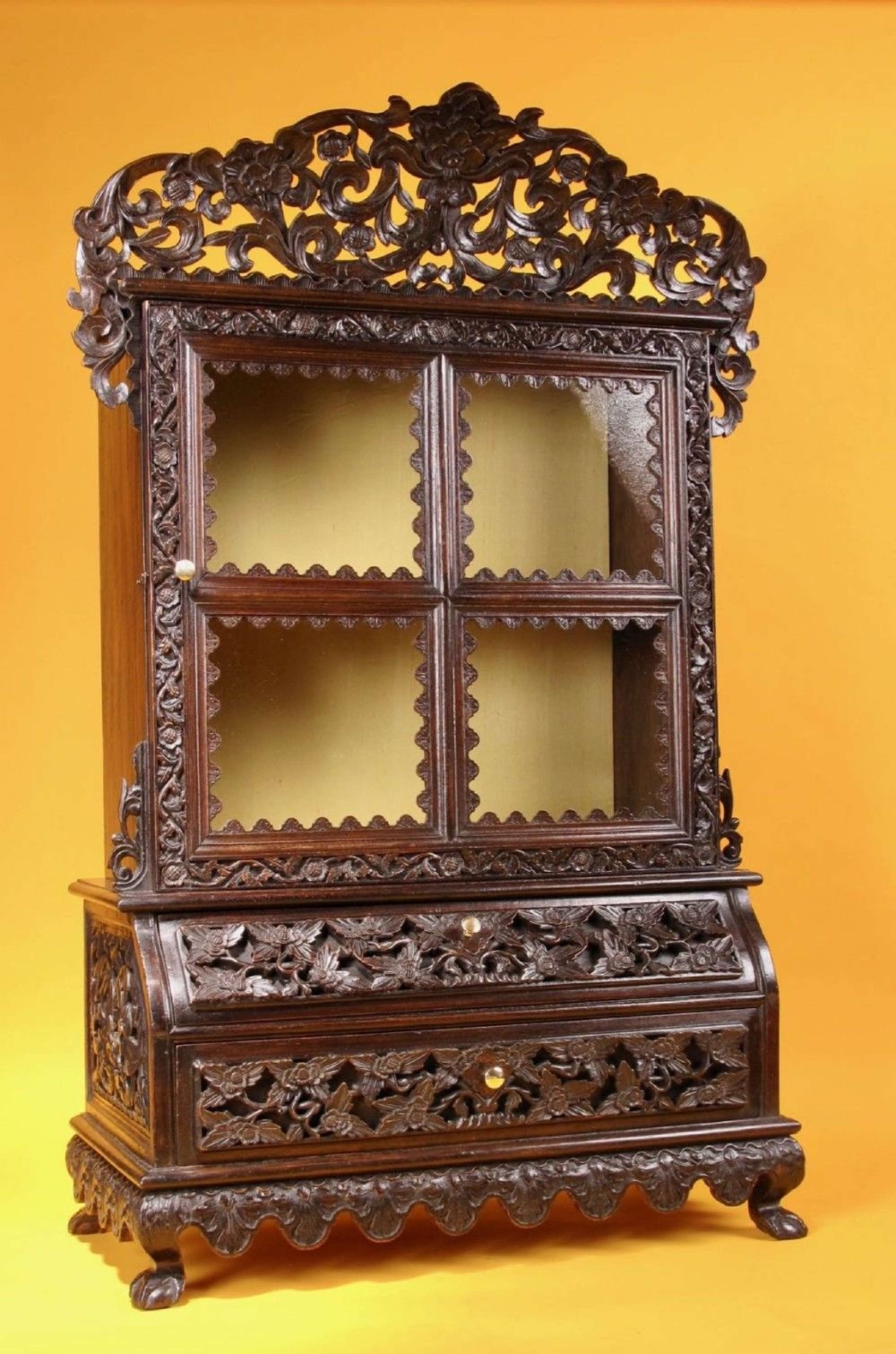 A 19th-century Anglo-Indian dark hardwood small display cabinet featuring intricate pierced foliate carvings, an arched pediment, a glazed four-pane door with scalloped moulding, and two carved drawers at the base.