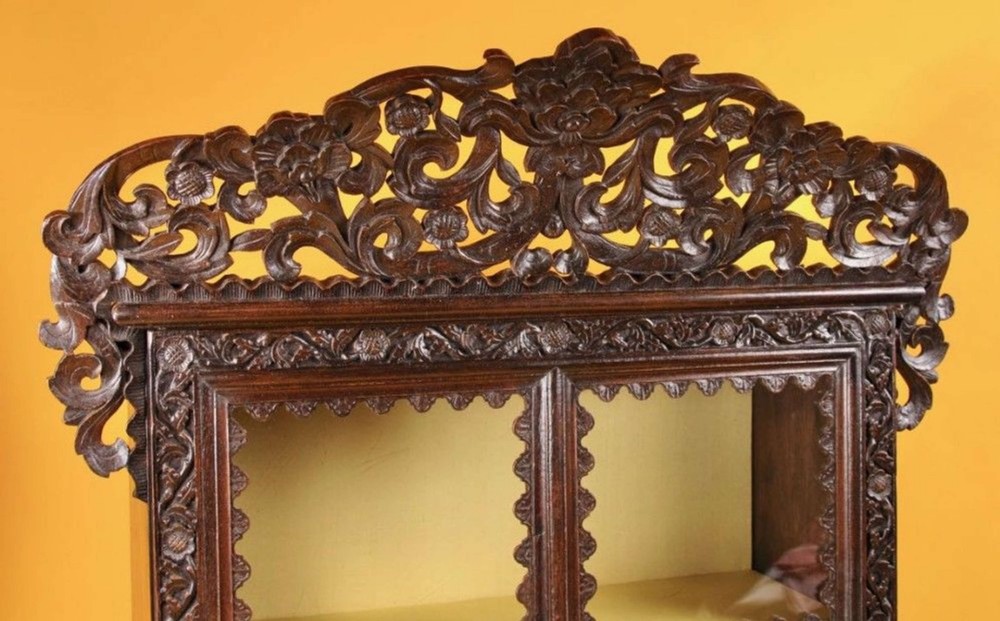 A 19th-century Anglo-Indian dark hardwood small display cabinet featuring intricate pierced foliate carvings, an arched pediment, a glazed four-pane door with scalloped moulding, and two carved drawers at the base. View X