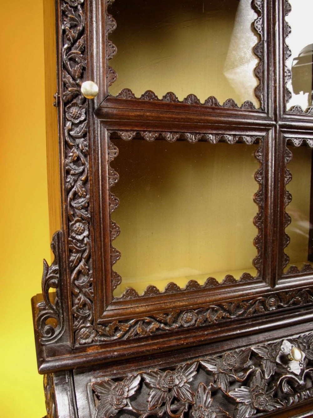 A 19th-century Anglo-Indian dark hardwood small display cabinet featuring intricate pierced foliate carvings, an arched pediment, a glazed four-pane door with scalloped moulding, and two carved drawers at the base. View XI