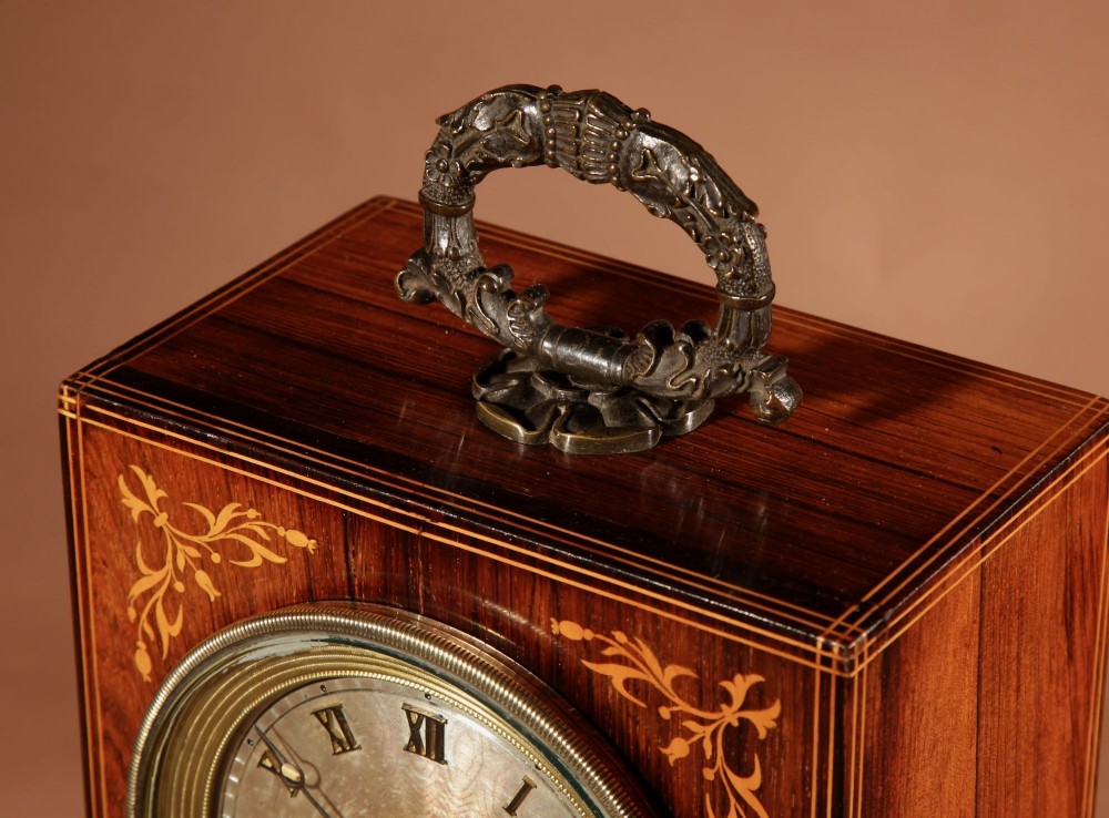 A rectangular Charles X dark rosewood mantel clock featuring intricate floral marquetry inlay, a circular silvered dial with Roman numerals, and a cast brass carrying handle on top. View VI