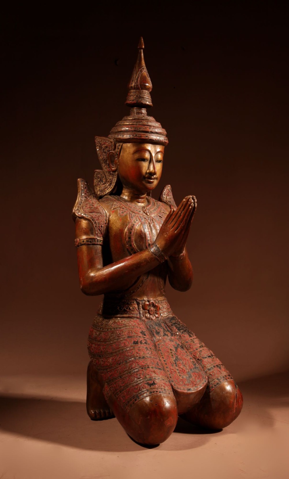 A carved wooden Thai Thepphanom guardian statue depicted kneeling with hands held in a prayer gesture, featuring a conical headdress and gilded red lacquered details. View II