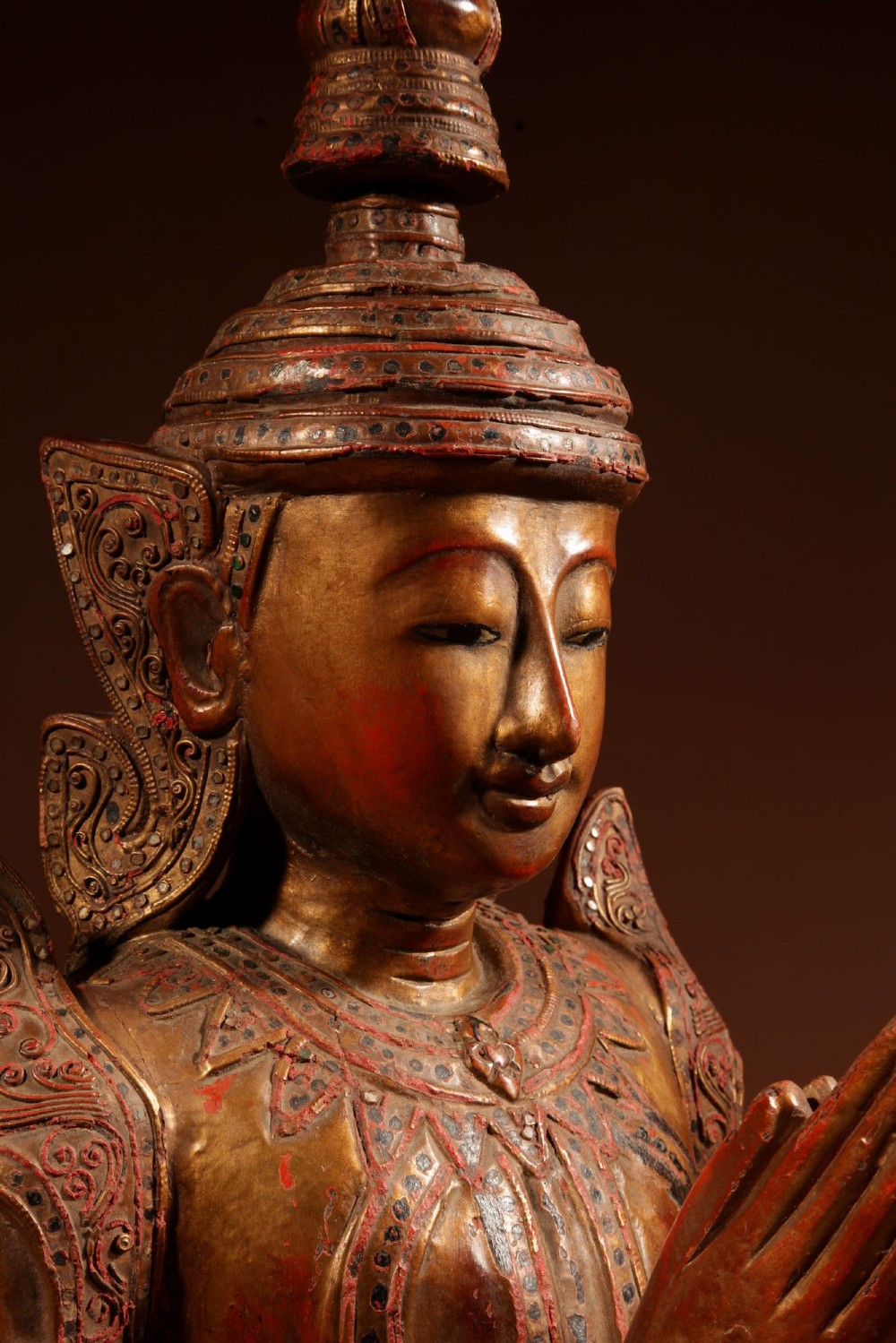 A carved wooden Thai Thepphanom guardian statue depicted kneeling with hands held in a prayer gesture, featuring a conical headdress and gilded red lacquered details. View III
