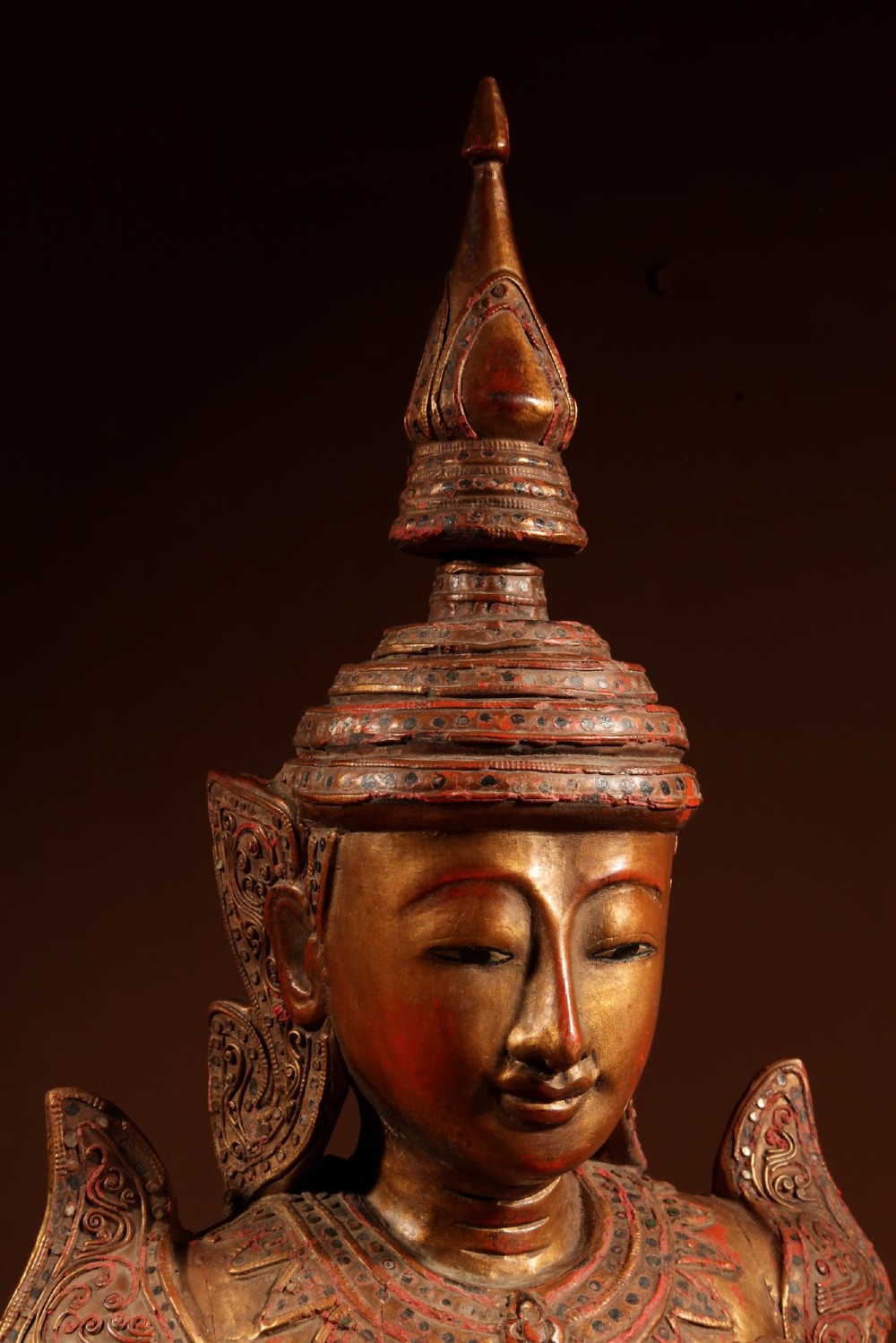 A carved wooden Thai Thepphanom guardian statue depicted kneeling with hands held in a prayer gesture, featuring a conical headdress and gilded red lacquered details. View IV