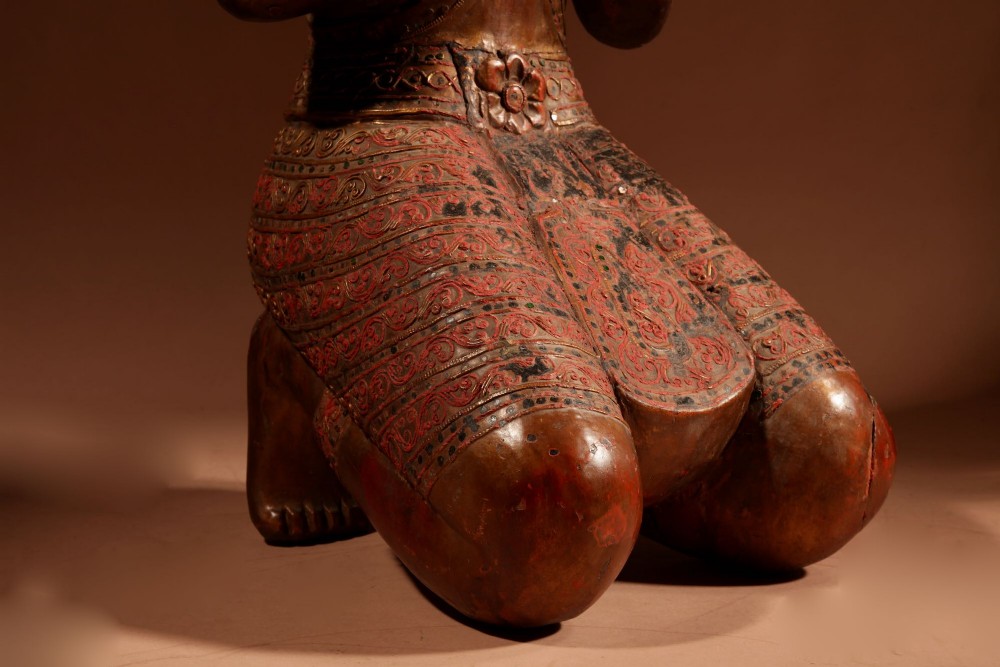 A carved wooden Thai Thepphanom guardian statue depicted kneeling with hands held in a prayer gesture, featuring a conical headdress and gilded red lacquered details. View V
