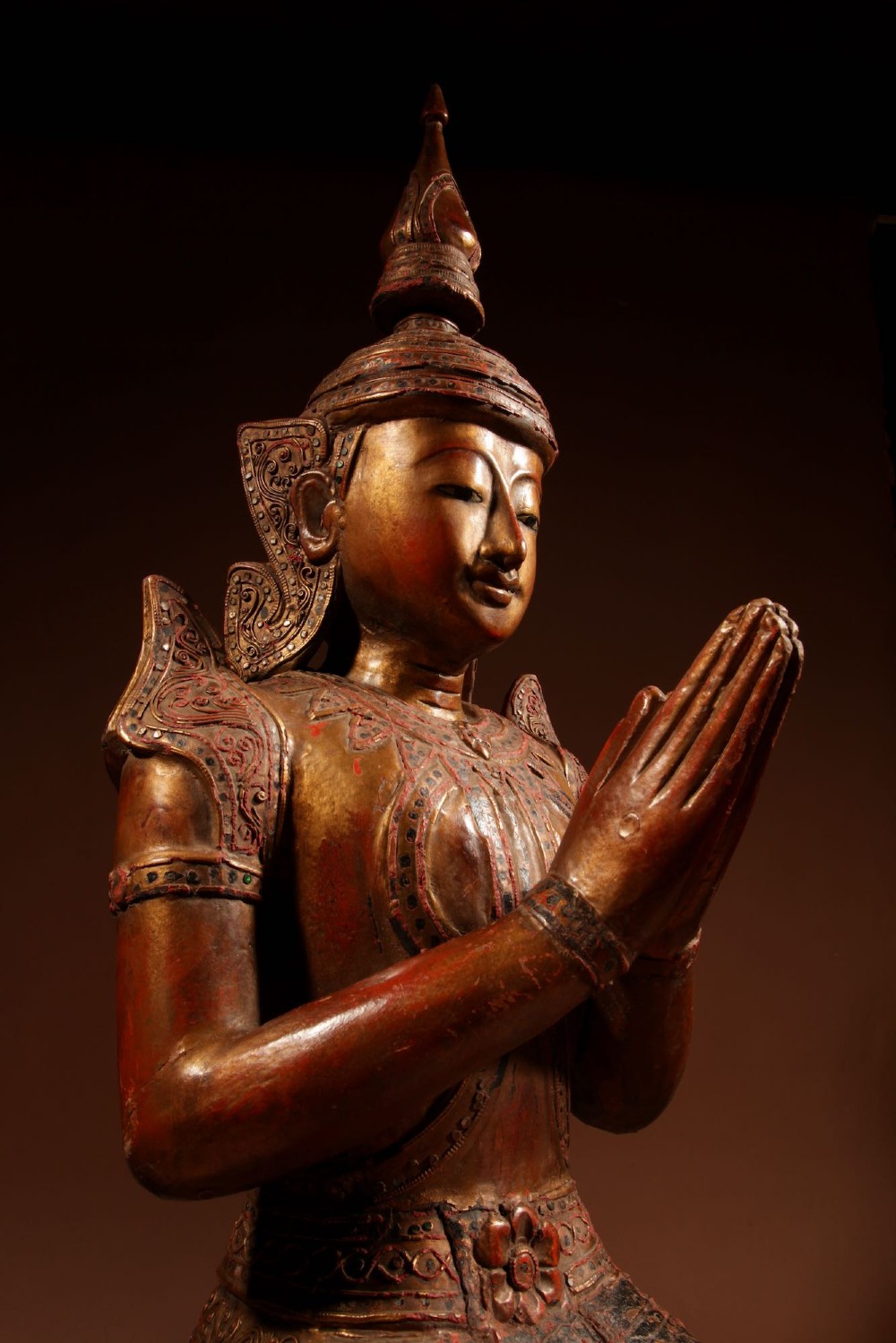 A carved wooden Thai Thepphanom guardian statue depicted kneeling with hands held in a prayer gesture, featuring a conical headdress and gilded red lacquered details. View VII