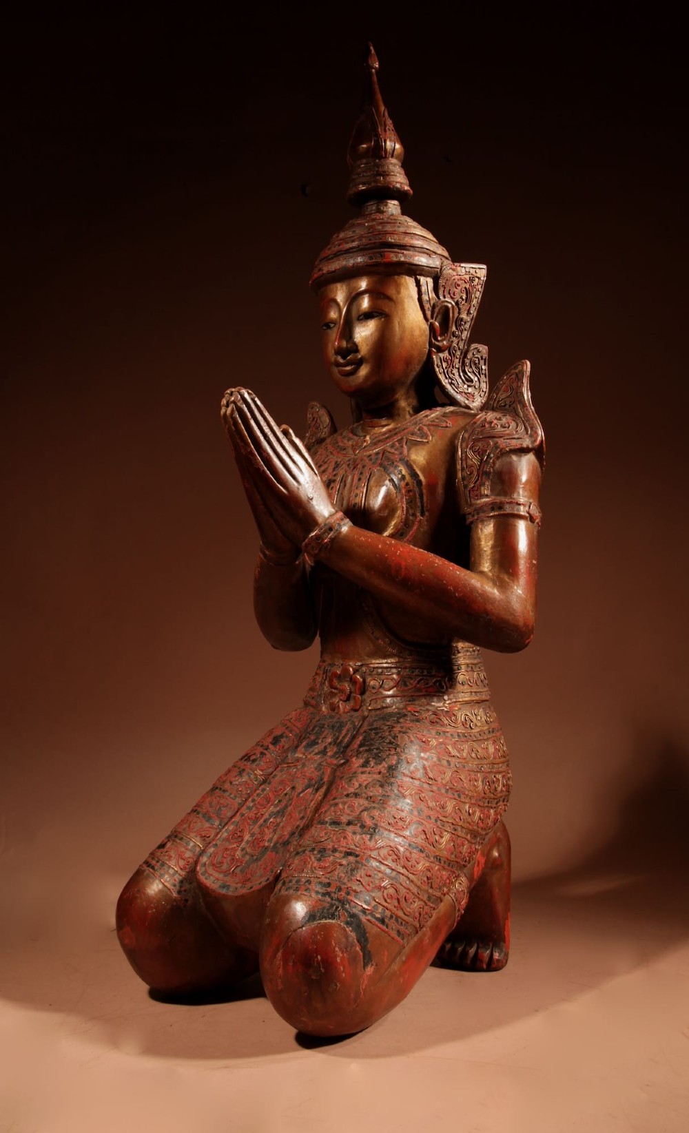 A carved wooden Thai Thepphanom guardian statue depicted kneeling with hands held in a prayer gesture, featuring a conical headdress and gilded red lacquered details. View VIII